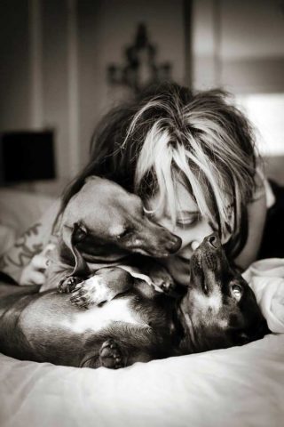 Pet family photo of a woman hugging her two Mini Dachshund on a bed.